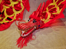 Tuotekuva Kotae Expon myymästä punaisesta 5 metriä pitkästä poi-lohikäärmeestä. Punainen poi-lohikäärme keltaisilla suomukuviolla ja kultakoristepisteillä. Lohikäärmeellä on pitkät rautalanka viikset, joiden päässä on kulkuset. sekä poi-lohikäärme beigellä pörröisellä taustalla.