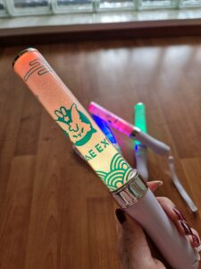 Kotae Expon valotikku, tuotekuva. Kuvan etualalla ihmisen käsi pitelee oranssilla valolla valaistua valkoinen valotikku. Valotikun valo-osassa on turkoosi perinteinen japanilainen aalto kaarikuvia alhaalla, keskellä Kotaen logo ja ylhäällä perinteinen japanilainen pilvi-kuvitus. Taustalla on ikkuna ja lattialla lisää Kotaen valotikkuja merenvihreällä ja magentan värisillä valoilla.